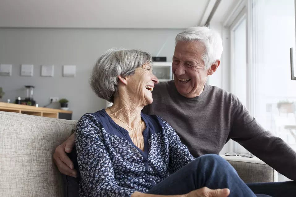 Senior couple in a couch happily laughing while looking into each others' eye / Couple de seniors dans un canapé, riant joyeusement en se regardant dans les yeux