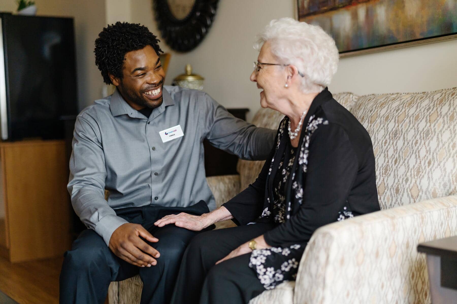 Chartwell care support staff smiling while sitting and chatting with the resident on couch 