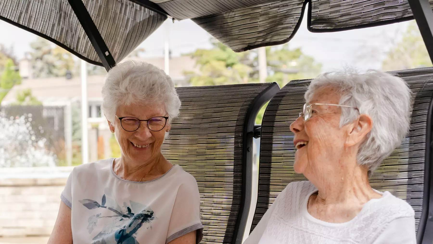 Two senior women are sitting on a swing chair and having fun conversation / Deux femmes âgées sont assises sur une balançoire et ont une conversation amusante