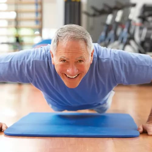 senior man wearing a blue shirt doing pushups on a blue mat on a wooden floor in a fitness room.