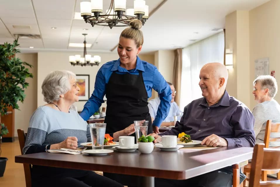 Chartwell Server having a pleasant conversation with the Senior couple at the dinning table / Le serveur de Chartwell a une conversation agréable avec le couple de personnes âgées à la table à manger