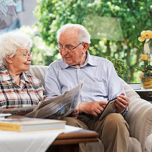 Senior couple is reading news from newspaper and a mobile device while sitting in a couch / Un couple de seniors, assis dans un canapé, lit les nouvelles dans un journal et sur un appareil mobile.