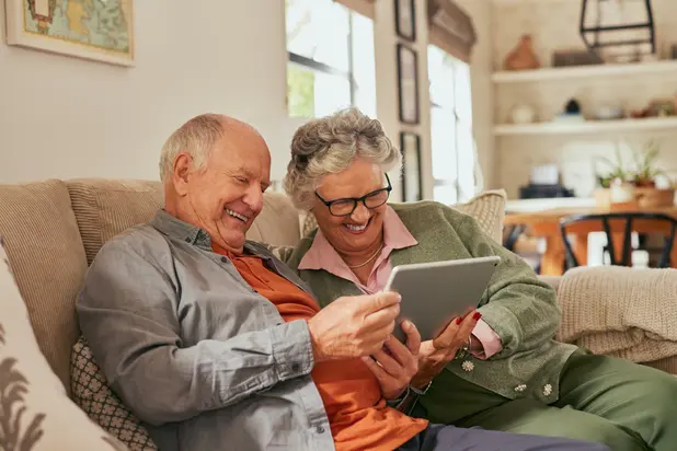 Laughing senior couple sitting on a couch using a digital tablet at home. Elder man and elderly woman exploring content and smiling together on a digital tablet. Carefree retired couple sharing a moment while using a touchscreen device with joy.