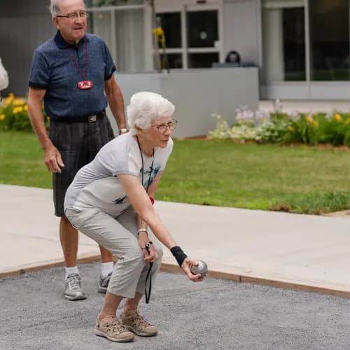 Senior lady crouching outside in a lawn bowling court, ready to throw a silver ball. There is a senior man standing off to the side behind her watching.