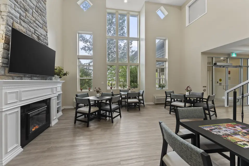 Bright lobby lounge with fireplace, large windows, and resident seating at Chartwell Royal Oak Village. / Salon du hall avec foyer, grandes fenêtres et sièges pour résidents au Chartwell Royal Oak Village.