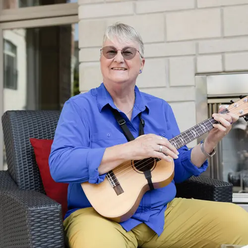 Outside sitting near the courtyard fireplace a senior lady in a bright blue shirt and fashionable mustard yellow pants is looking at the camera playing her ukelele.