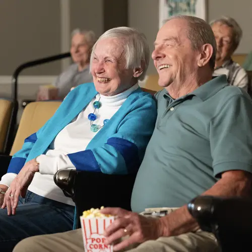 Couple sitting in a cinema looking towards a movie screen, smiling with man's arm around woman and the other hand holding a bag of popcorn.