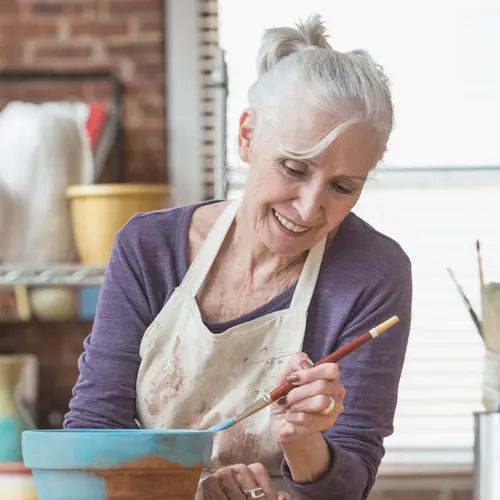 Senior woman in a studio painting a pot of cloy with a brush / Femme âgée dans un atelier peignant un pot de cloy au pinceau