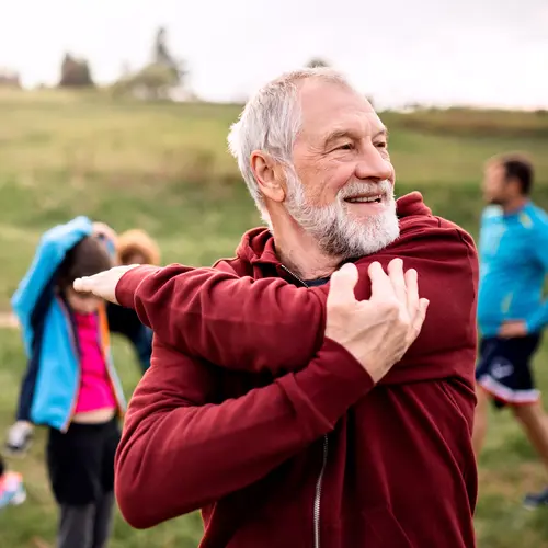 Senior man in wearing red hood, stretching out on a grass field / Homme âgé portant une cagoule rouge, s'étirant sur un champ d'herbe