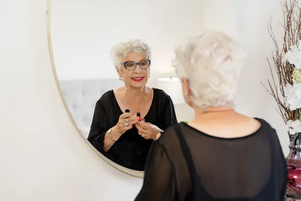 Senior woman is looking at an oval mirror while putting her red lipstick on / Une femme âgée regarde un miroir ovale tout en mettant son rouge à lèvres rouge.
