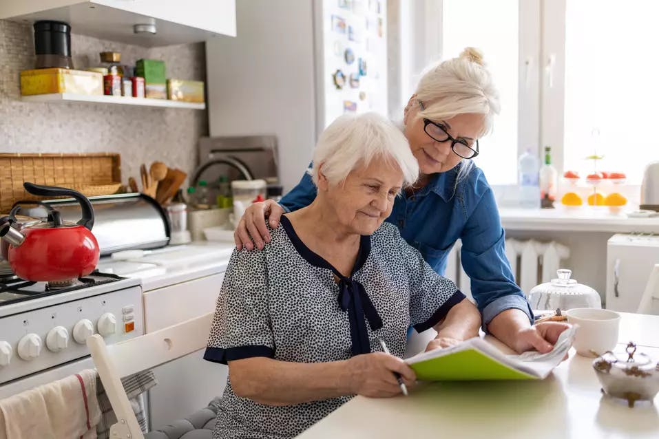 Senior woman and adult child in kitchen looking at notebook. Une femme âgée et un enfant adulte dans la cuisine regardant un cahier.