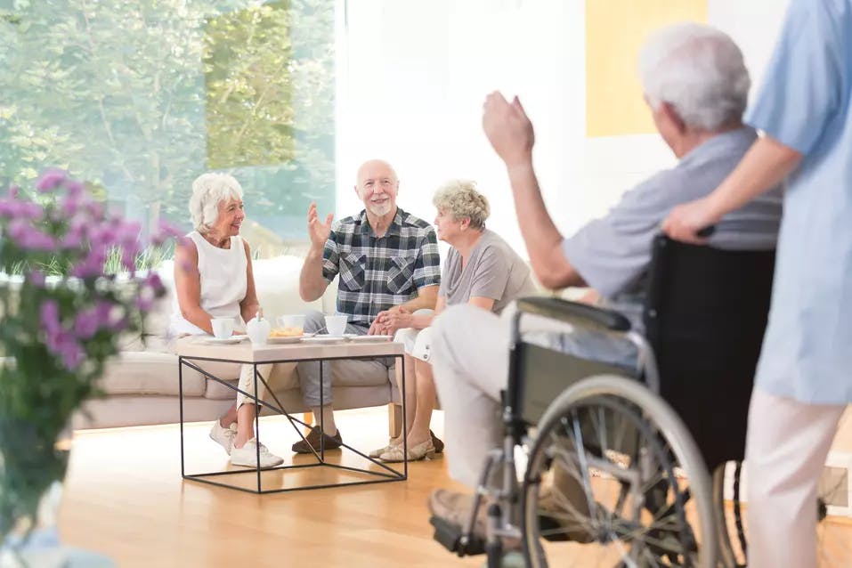Group of senior people welcoming a friend riding a wheelchair in a lounge / Groupe de personnes âgées accueillant un ami en fauteuil roulant dans un salon