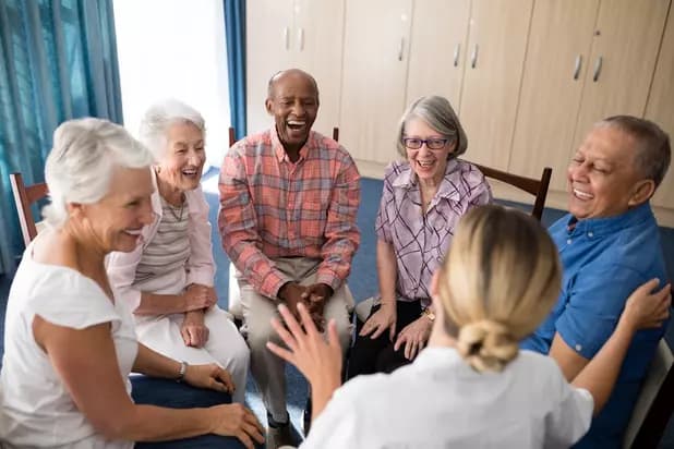 A group of seniors are sitting in a circle with a retirement residence staff and laughing happily / Un groupe de personnes âgées est assis en cercle avec le personnel d'une maison de retraite et rit joyeusement