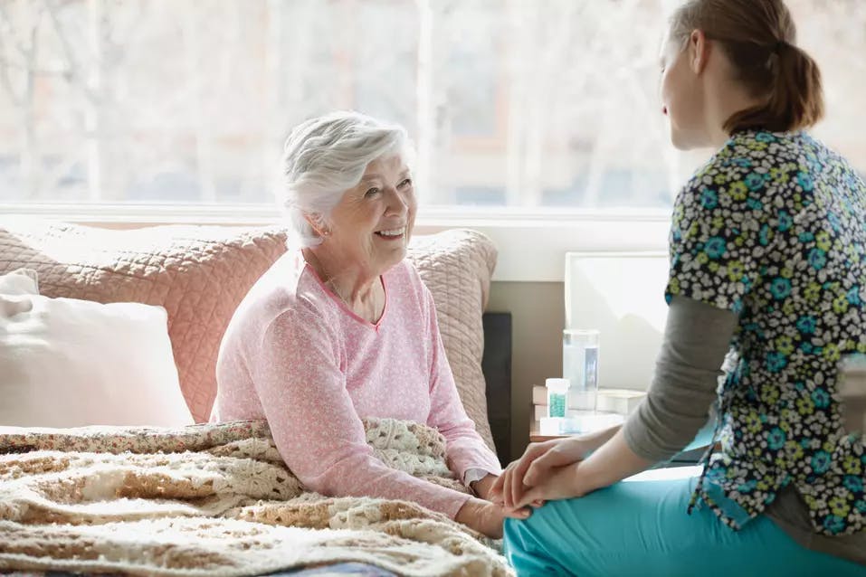 Senior woman sitting in bed smiling at her care worker sitting on a chair next to her